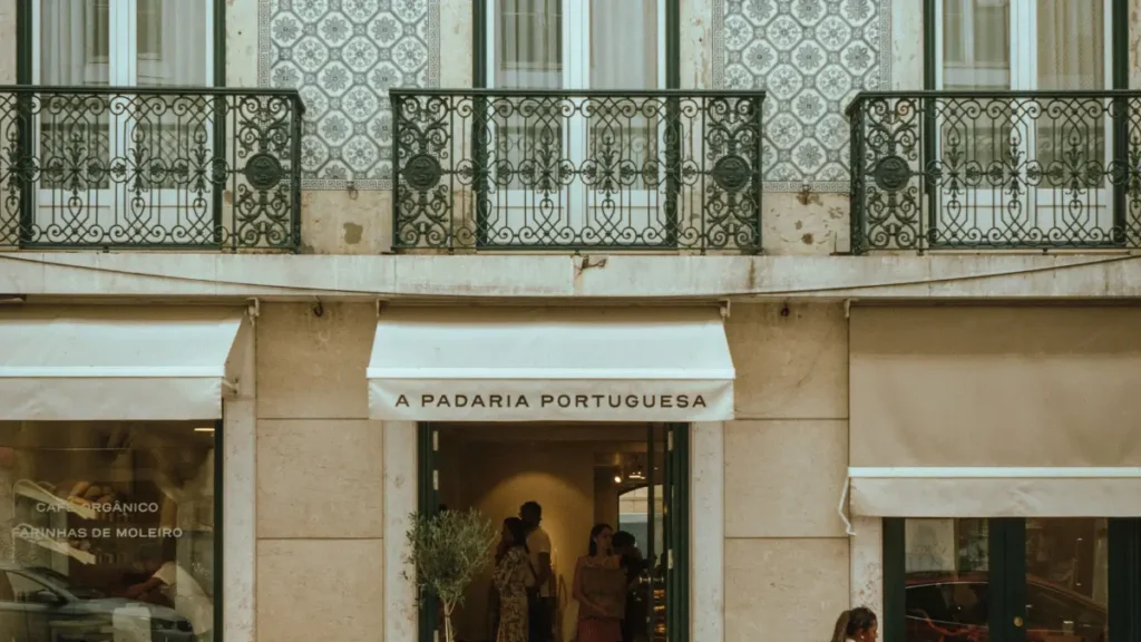 Fachada de pedra e toldo bege da A Padaria Portuguesa sob prédio com azulejos em Portugal.