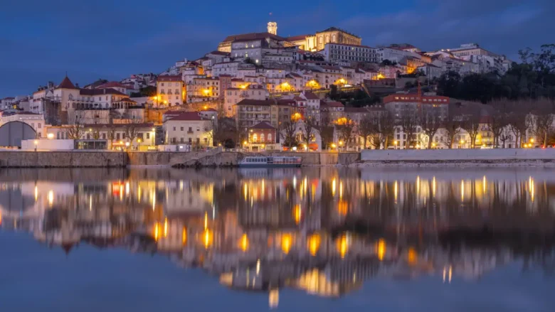 Vista noturna panorâmica da cidade de Coimbra com casario iluminado refletido no Rio Mondego.