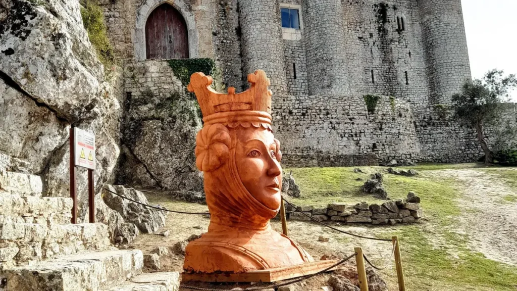 Escultura em terracota da cabeça de uma rainha em frente às torres de pedra do Castelo de Óbidos.