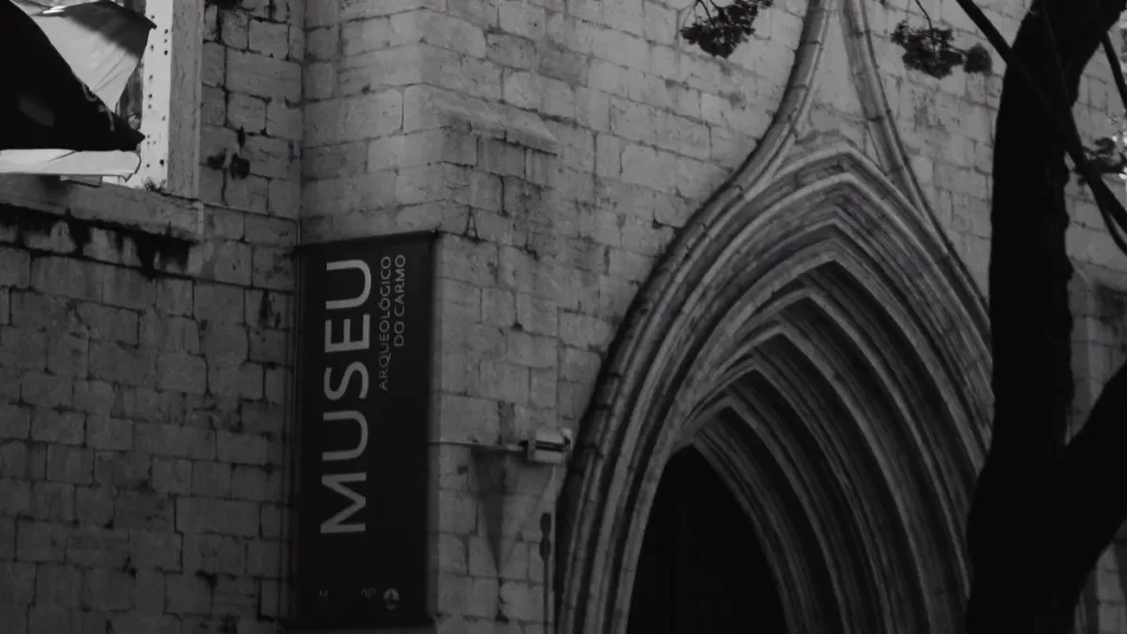 Fachada de pedra gótica do Museu Arqueológico do Carmo em Lisboa com portal em arco e cartaz visível, em foto preto e branco através de árvores.