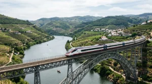 Trem de passageiros da CP cruzando uma ponte metálica sobre o Rio Douro cercado por vinhedos em Portugal.