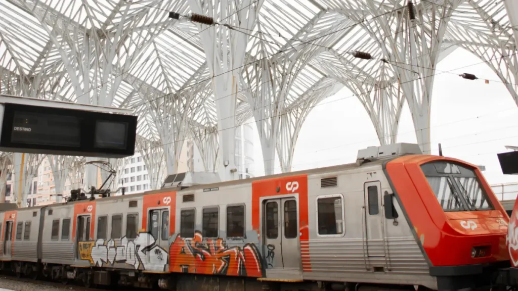 Trem da CP parado na plataforma sob a estrutura de aço da Estação Gare do Oriente em Lisboa.