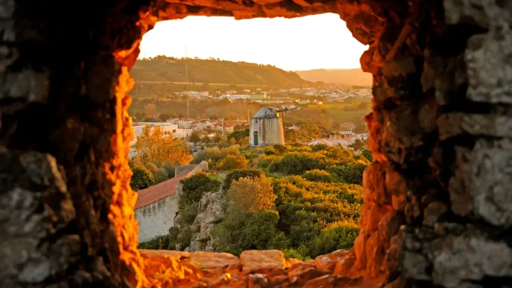 Vista emoldurada por janela de pedra da muralha de Óbidos mostrando moinho de vento e paisagem ao pôr do sol.